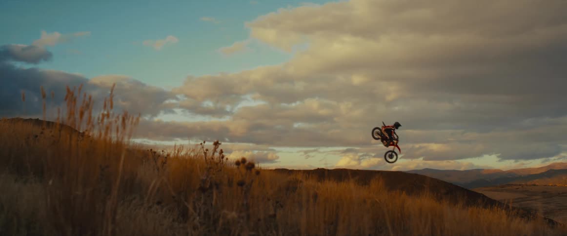 Epic slow motion footage captures a motocross rider catching big air off a dirt jump in a grassy, mountainous landscape at sunset