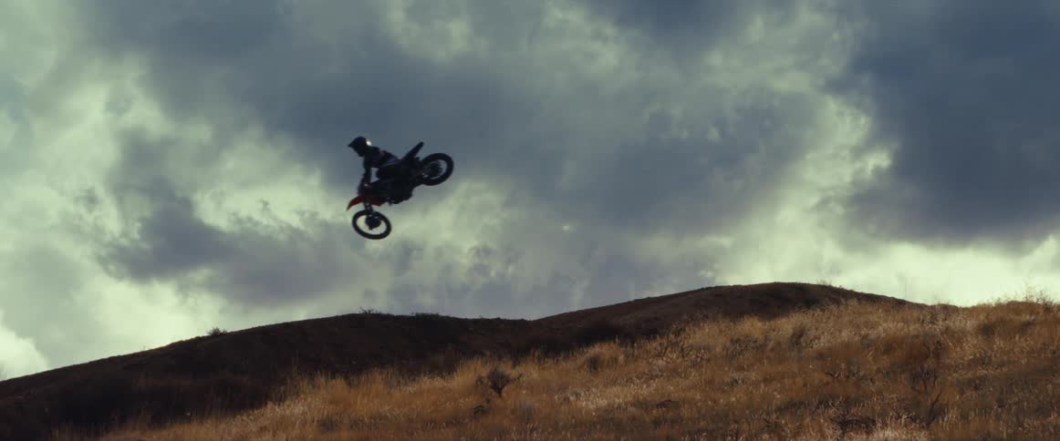 A motocross rider soars through the air in an epic slow-motion jump, silhouetted against a dramatic cloudy sky