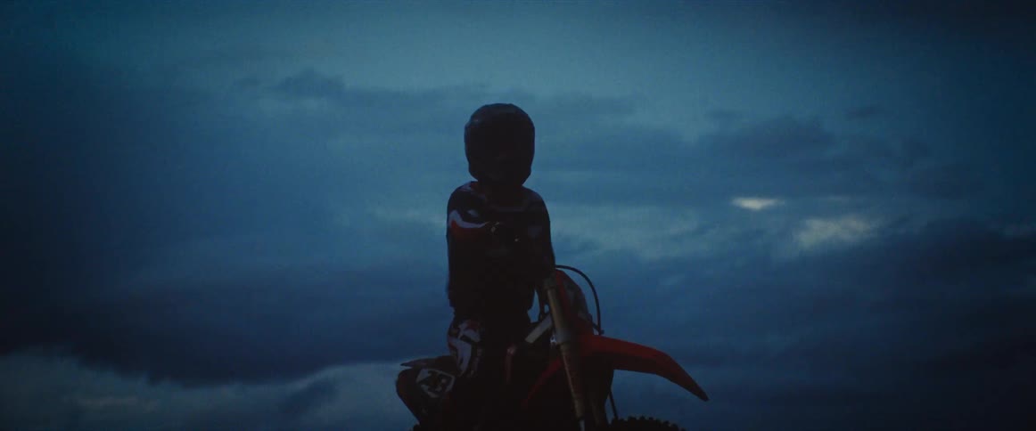 A silhouetted motocross rider sits on their dirt bike against a dramatic and dark cloudy sky at dusk