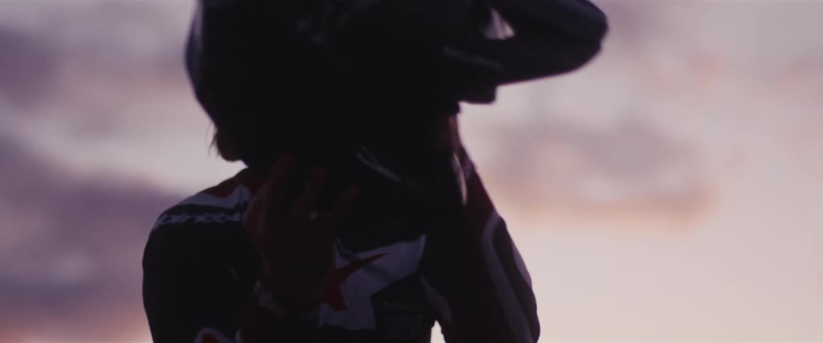 A silhouetted motocross rider prepares for a race by putting in his wireless earbuds and putting on his helmet at sunset