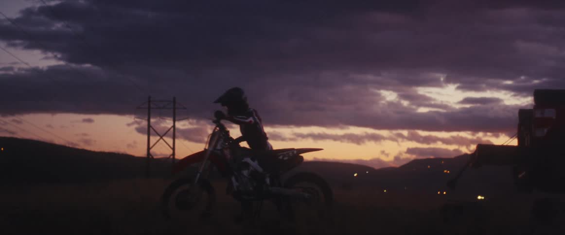 A motocross rider in silhouette slowly mounts their dirt bike against a dramatic sunset sky before riding off into the twilight