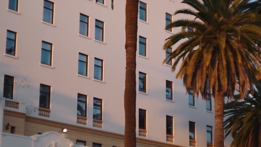 A slow pan reveals a classic white hotel with tall palm trees bathed in the warm light of a golden hour sunset