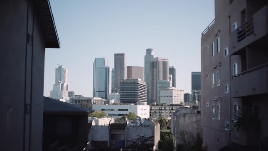 A slow, cinematic zoom towards the downtown Los Angeles skyline, viewed from between two shadowed apartment buildings on a clear day