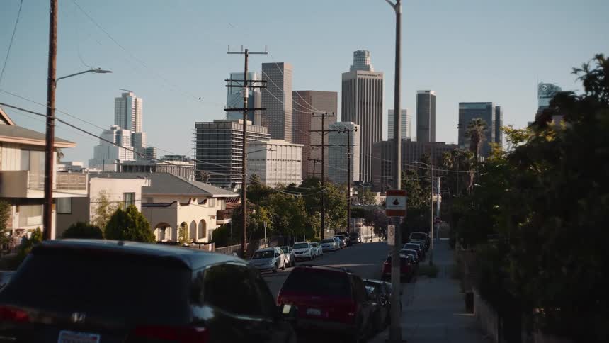 A slow push-in shot captures the downtown Los Angeles skyline from a residential street lined with parked cars and power lines