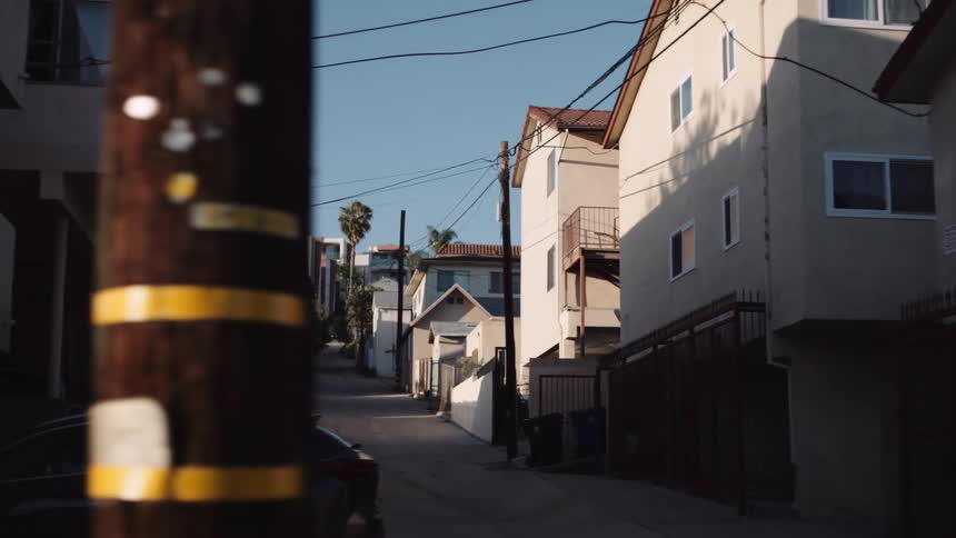 An observational slow pan from behind a parked truck reveals a quiet residential back alley on a sunny day