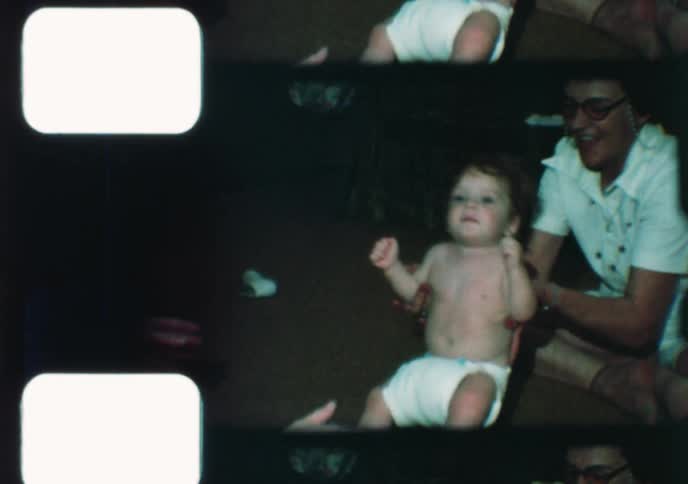 A vintage 8mm home movie shows a cute baby with curly hair taking its first wobbly steps with the help of its family