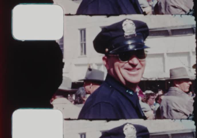 Vintage film captures a friendly police officer in sunglasses smiling while on duty at a public event