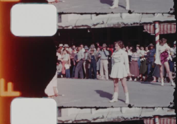 A vintage 8mm film scan shows a young majorette in uniform posing at a small town parade