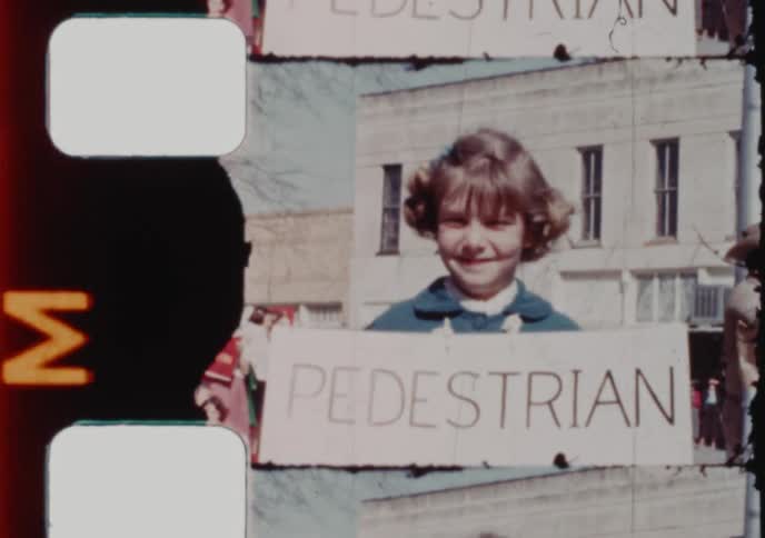 A proud little girl with pigtails smiles while holding a 'Pedestrian of the Week' sign in a vintage home movie
