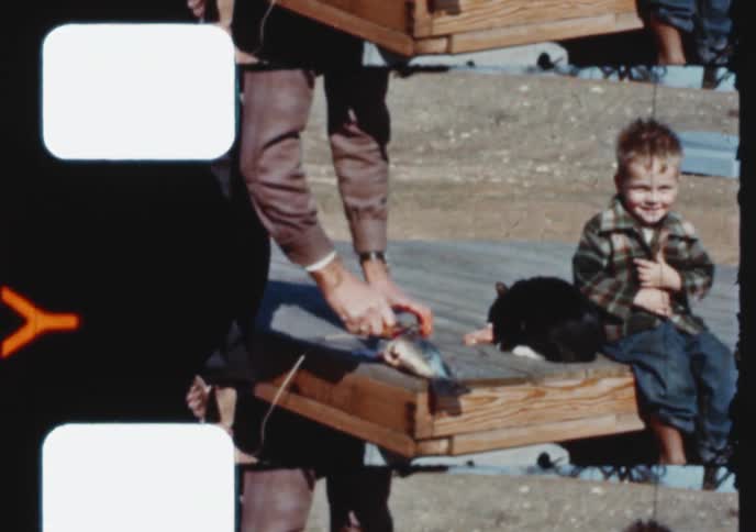 Vintage 8mm film shows a man cleaning a fish while a smiling little boy and a black cat watch
