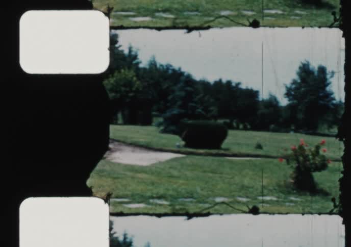 A vintage home movie captures a tilt down of a historic stone church, a smiling woman, and scenes from a sunny park