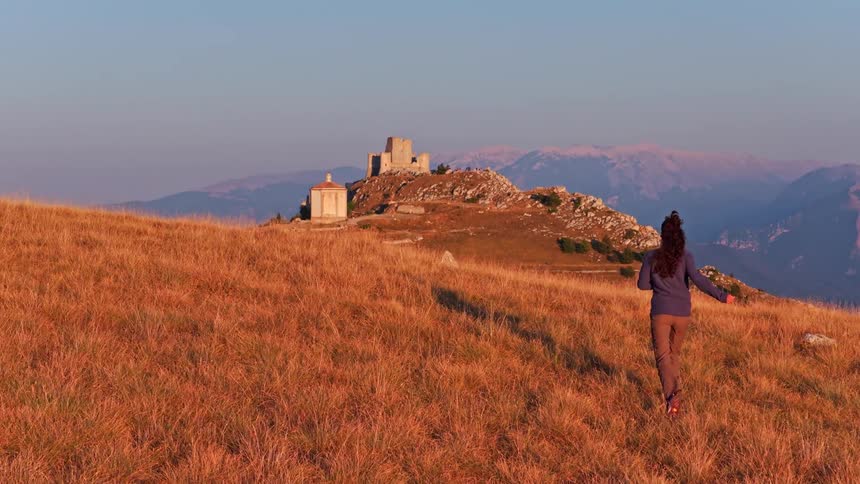 A woman runs through a golden, grassy mountain field towards the ancient Rocca Calascio fortress in Italy at sunset