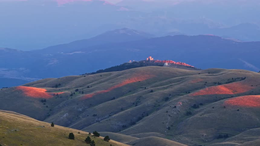 An aerial zoom out reveals a scenic mountain landscape with rolling hills and a distant castle catching the warm light of sunset