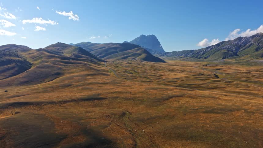 Static aerial view over the golden grasslands and rolling hills of Campo Imperatore valley, with the majestic Gran Sasso mountain under a clear blue sky in Italy