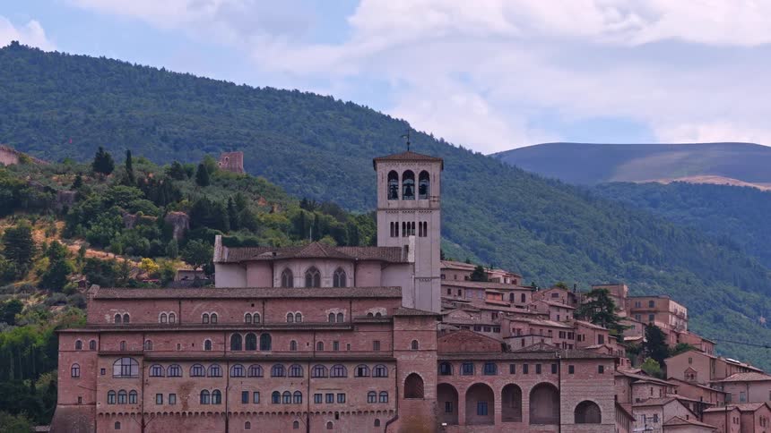 A slow zoom shot captures the magnificent Basilica of Saint Francis in the historic hillside town of Assisi, Italy, with lush green mountains in the background