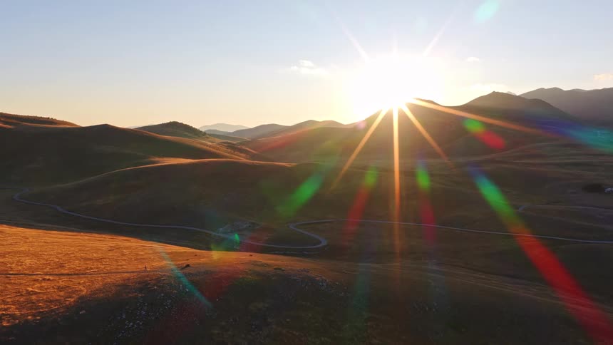 An epic aerial view shows the sun setting behind rolling hills, casting a golden glow and creating a vibrant lens flare over the vast mountain landscape