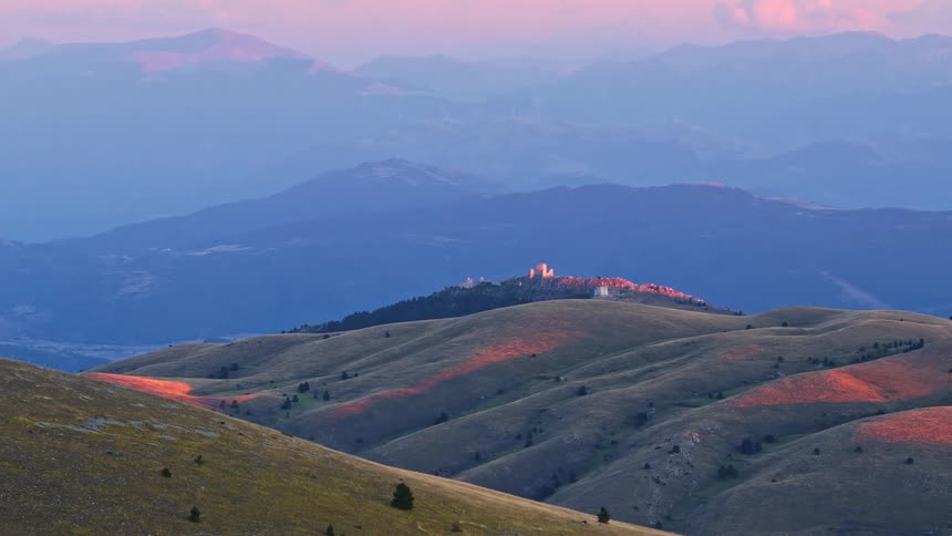 A panoramic view of the rolling hills of the Italian Apennine mountains with Rocca Calascio castle visible in the distance during a colorful sunset