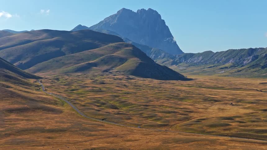 A stunning aerial view of a car driving on a winding road through the vast, golden plains of Campo Imperatore with the Gran Sasso mountain in the background