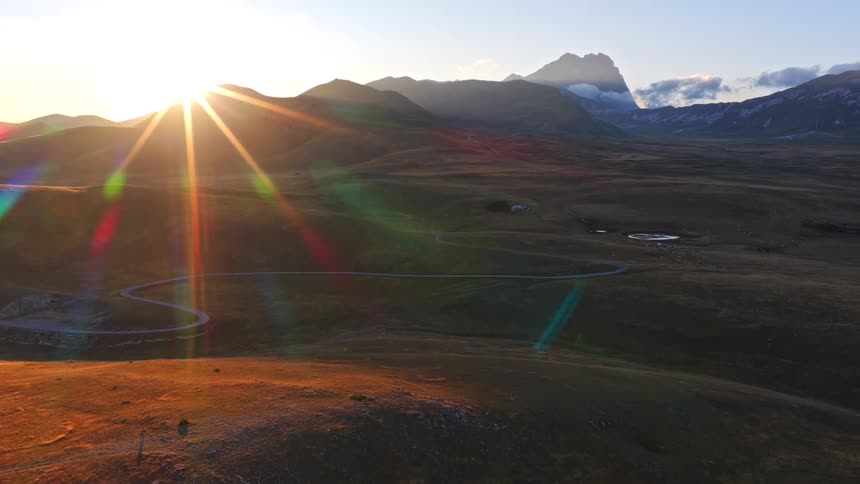 An aerial view shows the sun rising over a vast mountain landscape, creating a golden lens flare over a winding road