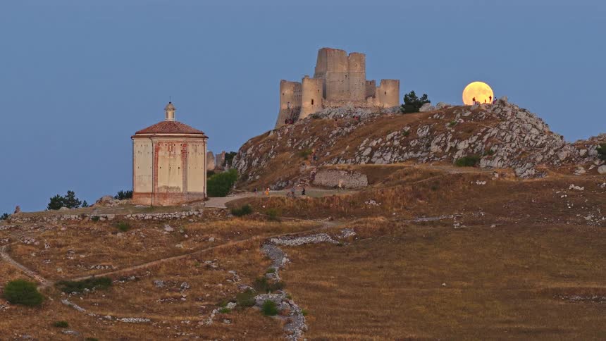A golden full moon rises in a time-lapse behind the ancient Rocca Calascio castle and Santa Maria della Pietà church in Abruzzo, Italy