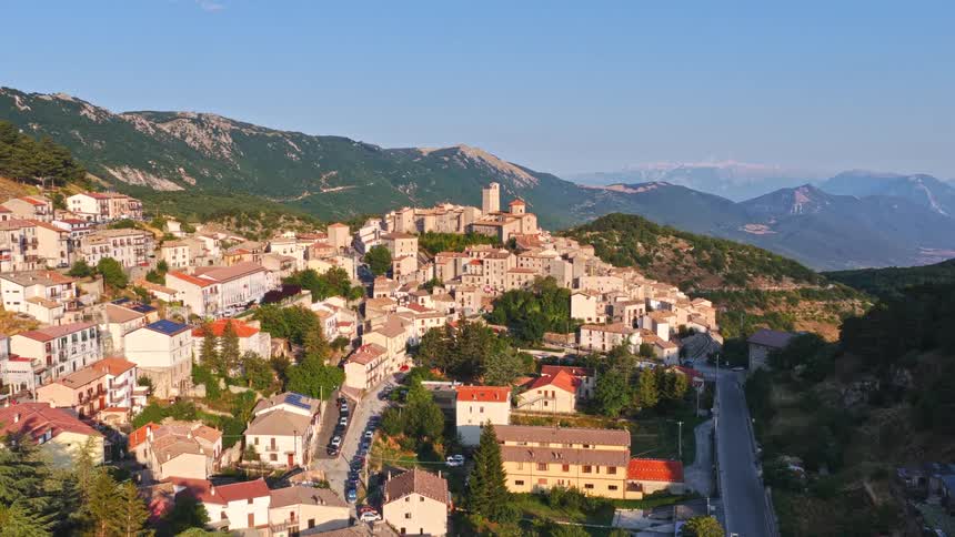 A stunning aerial view captures a picturesque Italian village nestled on a hillside, with a historic castle overlooking the town and majestic mountains in the background