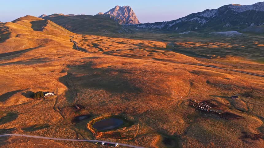 A stunning aerial tilt-up reveals a majestic mountain peak towering over a golden valley with sheep farms at sunrise