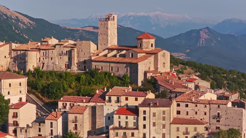 A picturesque zoom-in of an old Italian village perched on a green mountainside with a historic clock tower and church, set against a distant mountain range