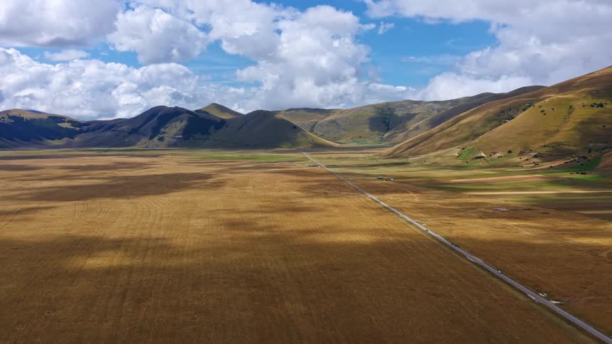 Sweeping aerial view of the vast, golden plains of Castelluccio di Norcia, with a straight road leading towards the mountains under a partly cloudy sky