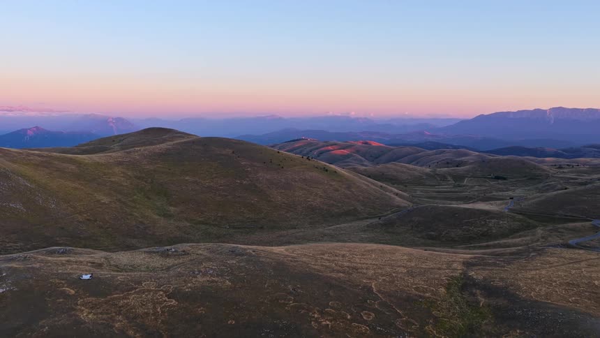 A serene aerial journey over vast golden hills and distant mountains reveals the moon rising during a beautiful sunset