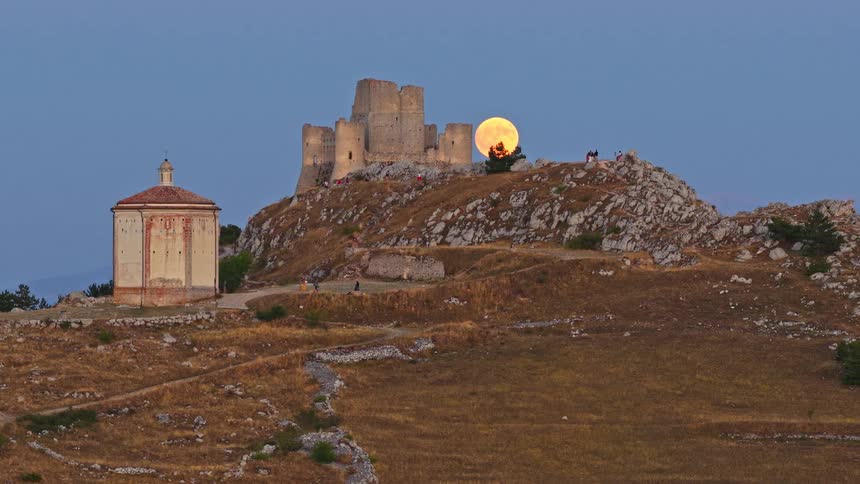 A stunning time-lapse of a full moon rising behind the ancient Rocca Calascio castle and the Santa Maria della Pietà chapel at dusk in Abruzzo, Italy
