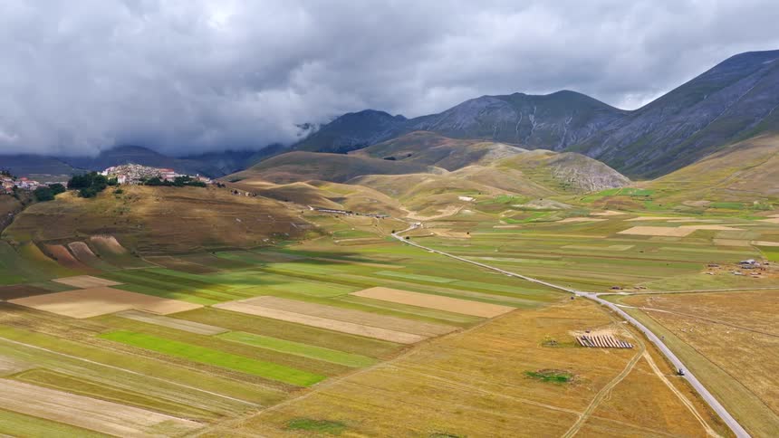 A breathtaking aerial view shows dramatic clouds passing over a vast valley of colorful patchwork fields nestled among mountains