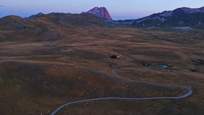 Breathtaking aerial view of a winding mountain road through a vast plateau as the sun rises, casting a warm glow on the distant peaks