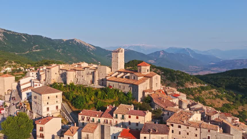 An aerial view slowly approaches the historic hilltop town of Casperia in Italy, showcasing its ancient stone clock tower and the vast mountain landscape beyond