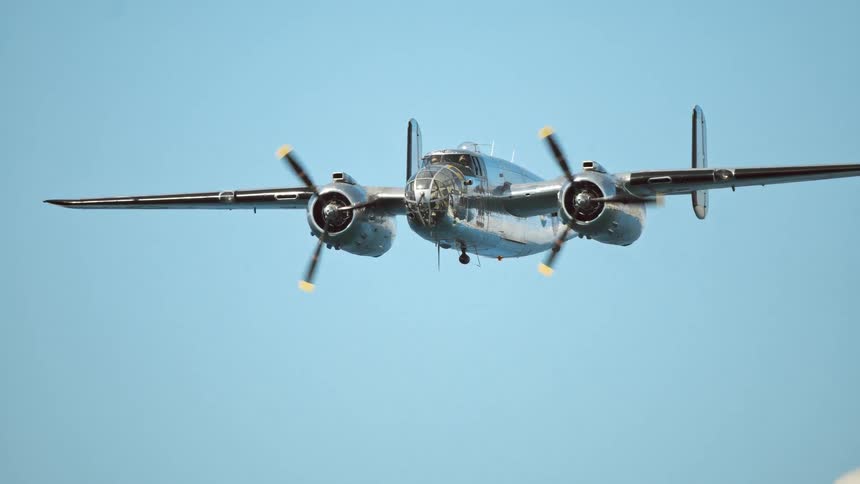 A vintage B-25 Mitchell bomber with a polished silver fuselage flies steadily through a clear blue sky in slow motion