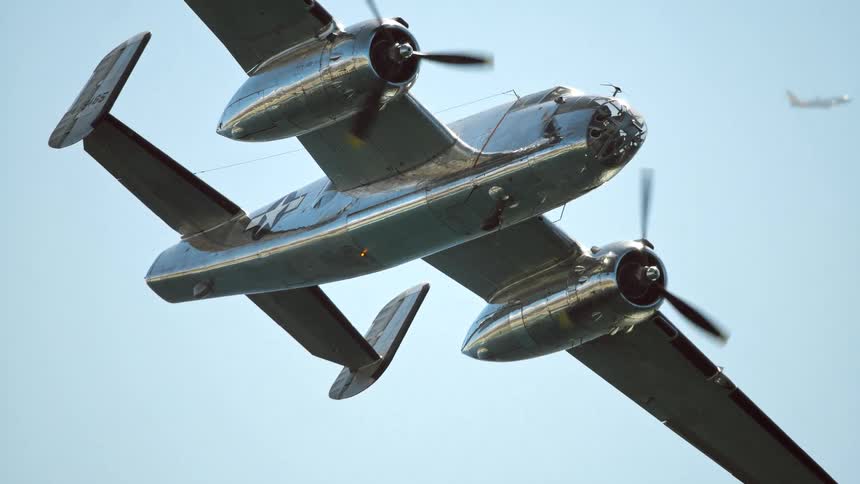 A polished chrome WWII twin-engine bomber soars majestically overhead in slow motion against a clear blue sky