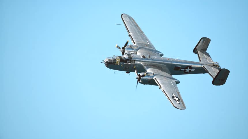 A classic World War II B-25 Mitchell bomber flies in slow motion against a clear blue sky