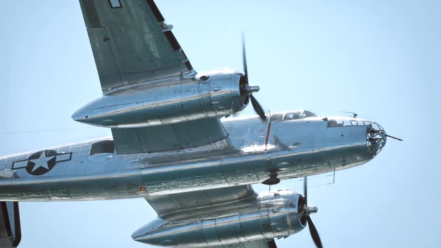 A gleaming silver World War II era bomber airplane flies in slow motion against a clear blue sky