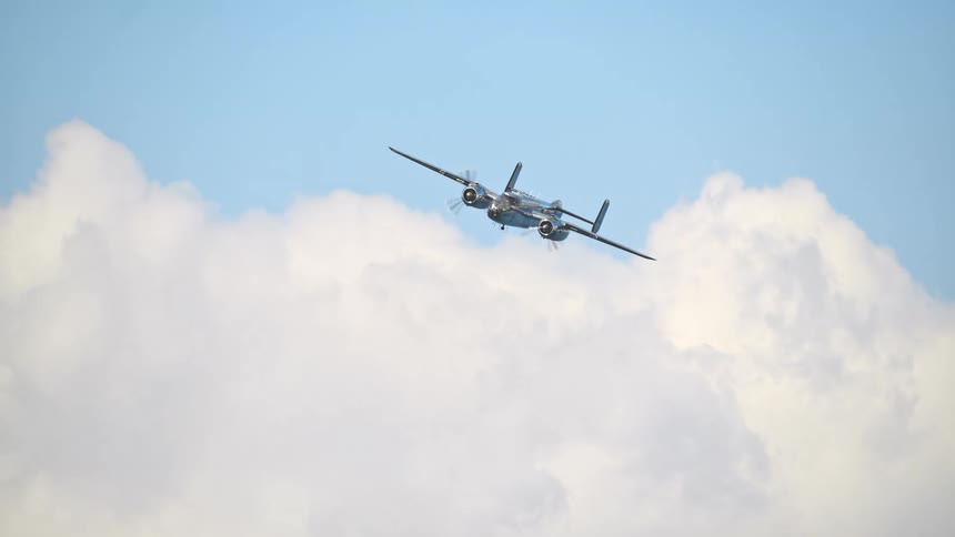 Slow motion footage shows a historic B-25 Mitchell bomber flying steadily towards the viewer against a sky of fluffy white clouds