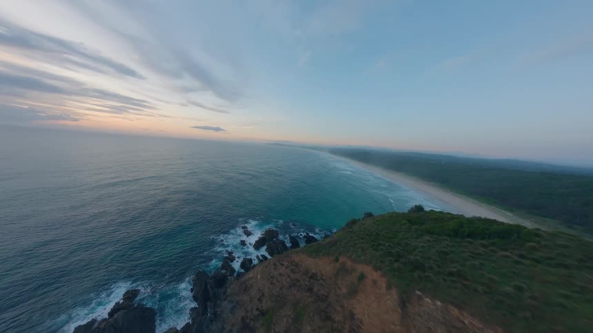 An FPV drone soars over a rugged, grassy headland at sunrise, revealing a stunning view of the vast ocean and a long, sandy coastline