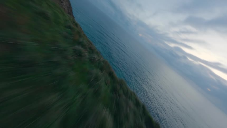 An exhilarating FPV drone flight dives down a lush, grassy cliff to follow the rugged coastline at dusk