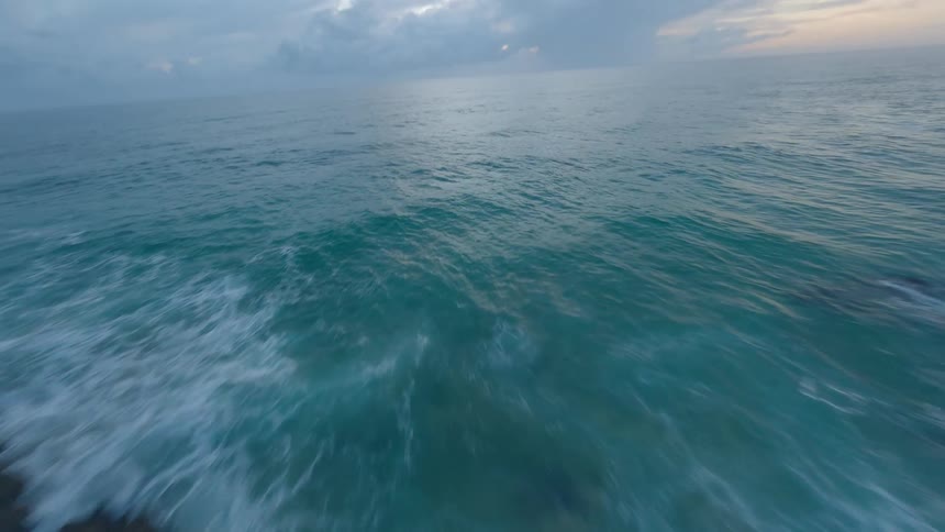 A fast-paced FPV drone shot descends a lush, rocky cliff and skims over the turbulent turquoise ocean at dusk