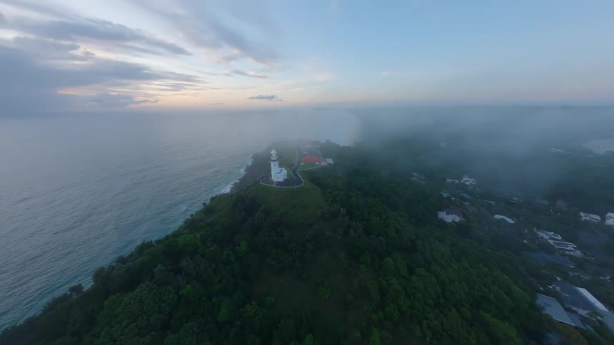 An ethereal aerial shot pushes in towards a majestic white lighthouse on a green coastal headland shrouded in mist at dawn