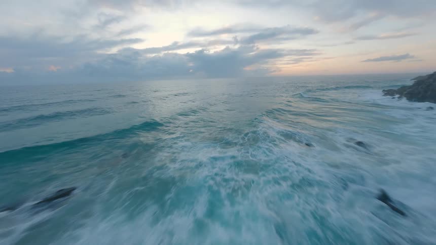 A dizzying FPV drone cliff dive down a rugged coastline transitions to a high-speed flight over the turquoise ocean waves at dusk