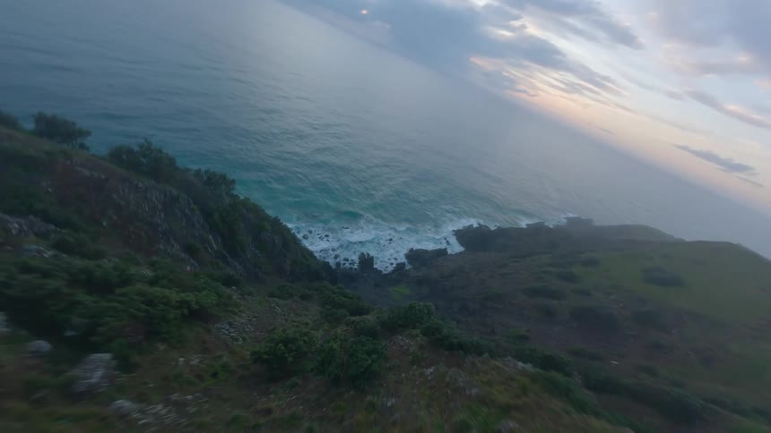 An FPV drone flies towards a foggy coastal lighthouse on a green cliff before diving dramatically towards the ocean waves below