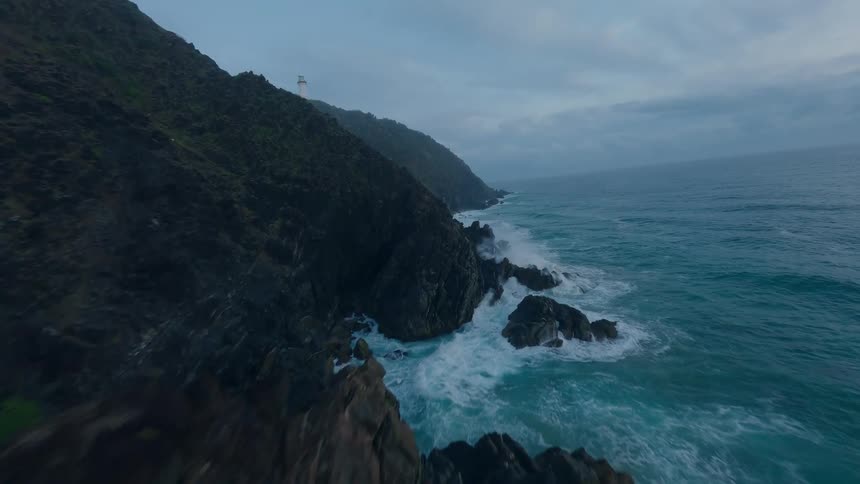 An exhilarating aerial view captures the raw power of the ocean as it crashes against rugged cliffs, with a lighthouse standing guard in the distance