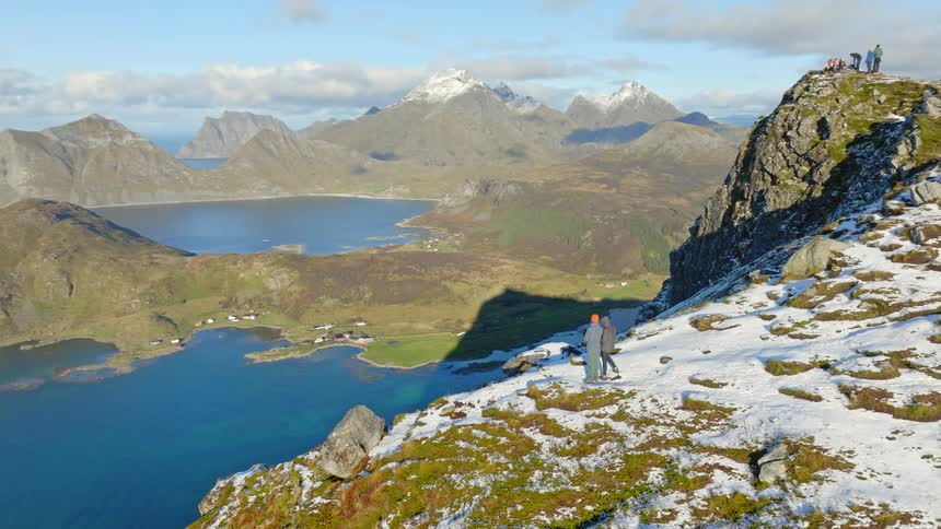 An aerial drone shot pans across a majestic snowy mountain peak revealing two hikers looking out at the stunning Norwegian fjords and ocean