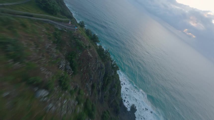 An FPV drone soars through the morning fog to a coastal lighthouse before dramatically diving down the cliffside to skim over the ocean waves