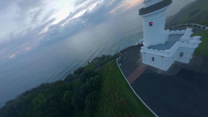 An FPV drone soars over a coastal peninsula, diving past a foggy lighthouse and down the cliffs to skim across the ocean waves at dawn
