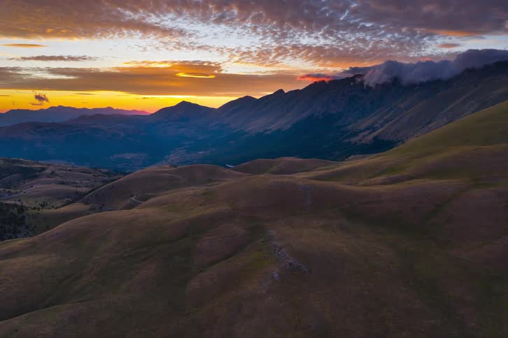 A stunning aerial time-lapse captures the sun setting behind majestic mountains, painting the sky with vibrant orange and purple hues over rolling hills