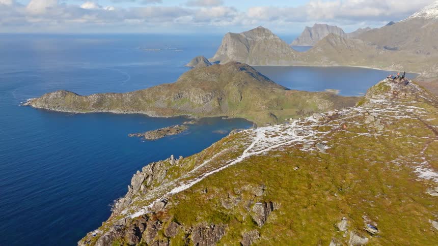 A dramatic aerial pullback reveals two hikers standing on a snowy mountain ridge overlooking a spectacular Norwegian fjord and ocean vista